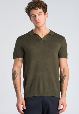 Giovane uomo con capelli ricci che indossa una polo a maniche corte in maglia verde oliva e pantaloni blu navy, con tatuaggi visibili su entrambi gli avambracci.