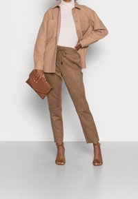Béžová košile-jacket z vlněné směsi s dvěma kapsami, spárovaná s tapered kalhotami z hnědé umělé semišové látky a hnědými lodičkami se zapínáním kolem kotníku, držící hnědou kabelku.
