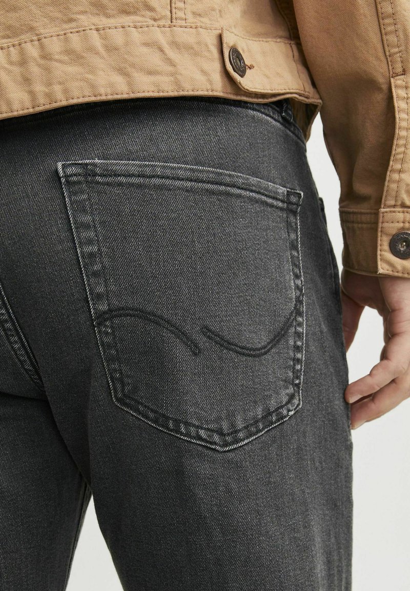 Dunkelgraue Jeans mit einer einzelnen Gesäßtasche, die ein gewundenes Nähtdesign aufweist. Hergestellt aus Denim, mit einer glatten Textur.