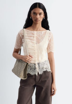 Giovane donna con lunghi capelli neri indossa una camicetta di pizzo crema trasparente, pantaloni scuri, orecchini a cerchio e porta una borsa a tracolla beige intrecciata.