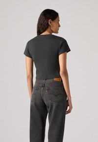 Schwarzes, gekürztes T-Shirt aus Baumwolle mit kurzen Ärmeln, kombiniert mit dunkelgrauen, hochgeschnittenen Jeans. Auffällige Taschenverarbeitung auf der Rückseite der Jeans.
