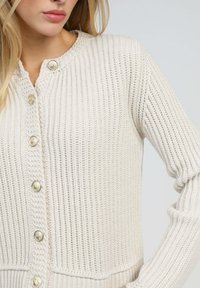 Femme portant un cardigan en tricot couleur crème avec des boutons décoratifs dorés, montré de la poitrine à la taille sur un fond clair.
