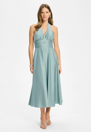 Femme portant une robe midi sans manches bleu sarcelle clair avec un col halter, une taille plissée et des talons argentés à brides, debout sur fond blanc.