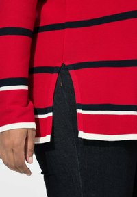 Sweter w czerwono-granatowe paski z rozciętym dołem i białymi akcentami, w połączeniu z ciemnymi dżinsami. Zbliżenie na rękaw i dłoń.