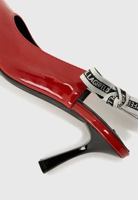 Zapato de tacón alto de charol rojo con lados abiertos, tacón negro y una correa blanca con texto negro. Textura suave, diseño elegante.
