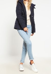 Mørkeblå puffjakke med pelsfor i hætten, lynlås foran og quiltet design, parret med lys blå rullede jeans og hvide sneakers.