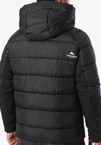 Homme portant une doudoune noire avec capuche, montrant son dos avec le logo "Helvetica" et un motif de montagne en haut à droite.