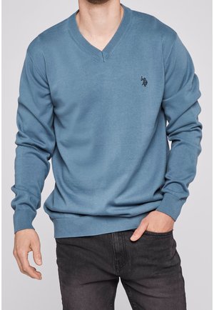 Homme portant un pull bleu à col en V et manches longues avec un petit logo sombre sur la poitrine, associé à un jean noir, une main dans la poche.