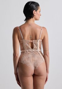 Intimo in un morbido beige, con materiale trasparente, dettagli in pizzo, schiena a occhiello e spalline regolabili. Motivo di pizzo intricato sul fondo.