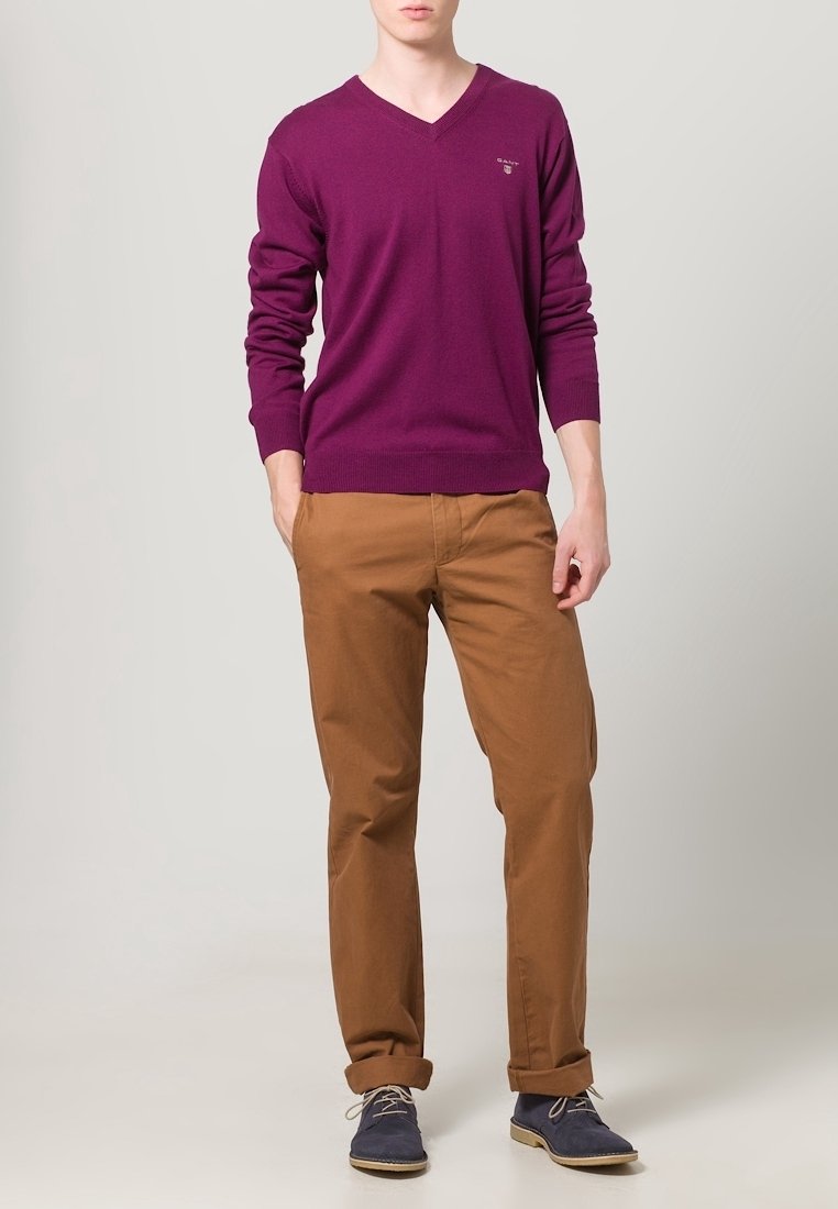 V-Ausschnitt lila Pullover, braune Chinos und dunkelblaue Schuhe. Der Pullover hat lange Ärmel; die Hose ist im geraden Schnitt mit hochgekrempelten Säumen.