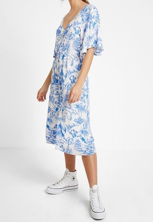 Mujer con vestido midi blanco con estampado tropical azul y mangas con vuelo, combinada con zapatillas blancas altas sobre un fondo liso.