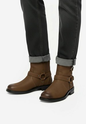 Brązowe skórzane botki za kostkę z metalowym pierścieniem i paskiem, noszone z czarnymi podwiniętymi dżinsami.