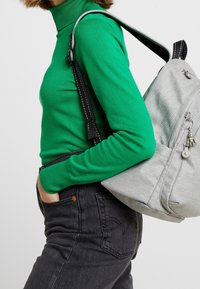 Zelený rolák, šedý batoh s texturovanou látkou a detaily na zip, v kombinaci s tmavými džíny, ukazující design ležérního oblečení.