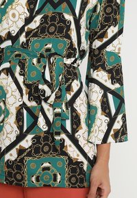 Top a fantasia verde, nera e bianca con disegni di catene dorate, cintura annodata in vita e maniche a campana di tre quarti, indossato da una persona con pantaloni marroni.