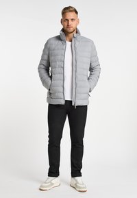 Giacca a vento grigia con cuciture verticali, colletto alto, chiusura con zip, due tasche laterali, indossata sopra una maglietta bianca e pantaloni neri.