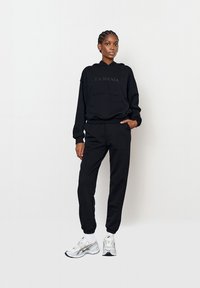 Hanorac negru cu logo-ul "LA MANIA", buzunar frontal, asortat cu pantalonii de trening de aceeasi culoare. Modelul poartă pantofi sport albi cu accente argintii.