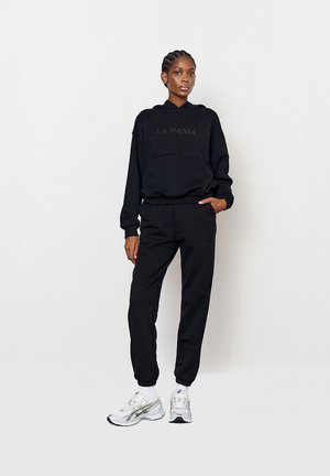Černá mikina s kapucí s logem "LA MANIA", přední kapsa, v kombinaci se stejnými tepláky. Model nosí bílé sportovní boty se stříbrnými akcenty.