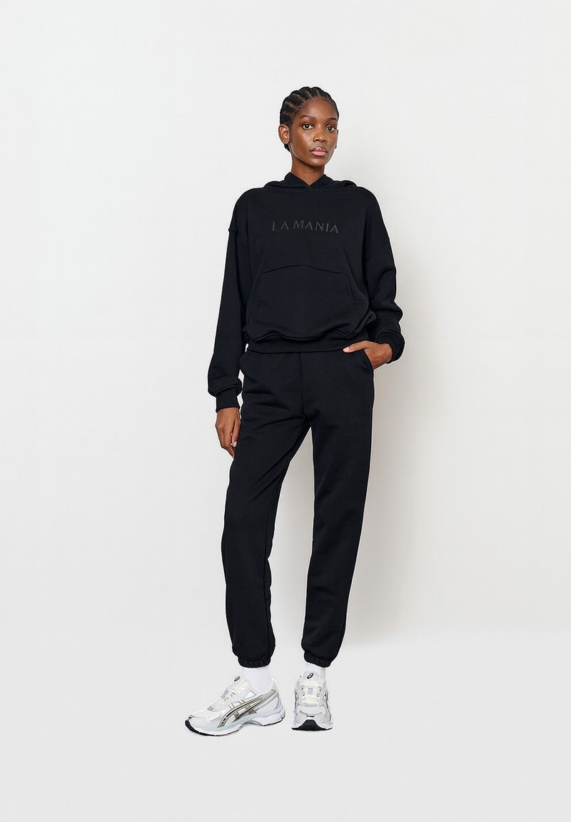 Hanorac negru cu logo-ul "LA MANIA", buzunar frontal, asortat cu pantalonii de trening de aceeasi culoare. Modelul poartă pantofi sport albi cu accente argintii.