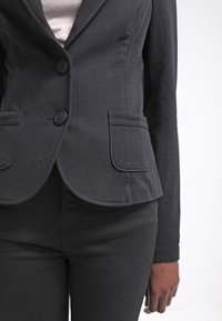 Femme portant un blazer noir cintré avec deux boutons et des poches à l'avant, associé à un pantalon noir, debout devant un fond blanc.