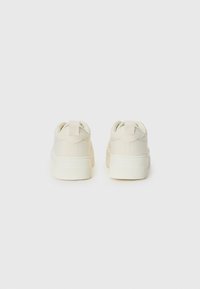 Pair of rear-view light beige sneakers with a textured canvas upper, padded collar, pull tab, and a thick white rubber sole.