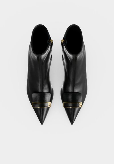 Paire de bottines noires à bout pointu avec un détail de boucle rectangulaire dorée sur le dessus, vue de dessus sur fond blanc.