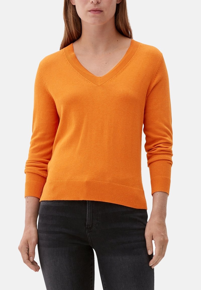 Femme portant un pull orange vif à col en V avec manches longues et un jean noir, debout sur un fond clair uni.