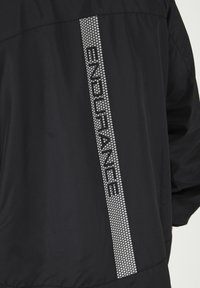 Veste noire avec un texte réfléchissant argenté "ENDURANCE" le long d'une rayure pointillée verticale sur le côté gauche. Texture de tissu lisse.