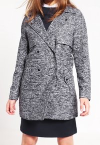 Cappotto grigio strutturato doppiopetto con rever a punta, due tasche laterali e bottoni neri. Indossato sopra un vestito scuro. Finitura in tessuto liscia.