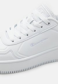 Hvite sneakers laget av syntetisk lær med flate snører, avrundet tupp og preget logo-detalj på siden. Teksturert såle.
