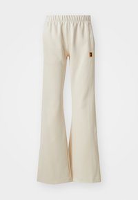 Pantalones acampanados beige, hechos de una tela suave con una cintura elástica y un bolsillo lateral. Presenta un pequeño parche con el logo en la cadera.