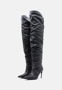 Botas pretas até ao joelho feitas de couro macio e enrugado, com bico fino e salto agulha. Textura lisa e ajuste justo.