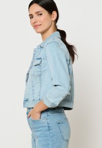 Hele sinine teksajakk, millel on lühike disain, nööbid, kaks rinnataskut ja üles keeratud varrukad, millele on sobitatud vastavad teksad.