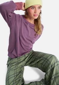 Sudadera morada con rayas de punto rosa en los puños, presenta un logotipo floral. Pantalones de pata de elefante verdes de cuadros con líneas de rejilla negras. Gorro amarillo.