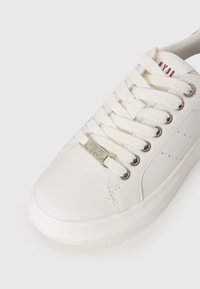 Hvide lærreds-sneakers med rund tå, flade snørebånd, metaløjer, tekstureret sål og et metal-logo-ornament på siden.