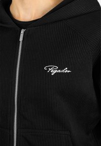 Čierna pletená mikina na zips s kapucňou, so malým bielym vyšitým logom na ľavej strane a prednými vreckami.