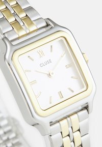 Silber- und goldfarbene Edelstahl-Uhr mit quadratischem Ziffernblatt, weißem Ziffernblatt, goldenen Akzenten und minimalistischen Stundenmarkierungen. Cluse-Marke angezeigt.