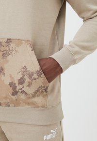 Sudadera color caqui con un bolsillo estampado en camuflaje, tela texturizada, puños acanalados y un logo blanco en la parte inferior izquierda.