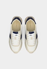 Ζευγάρι λευκά και μπεζ αθλητικά παπούτσια με ναυτικό μπλε λεπτομέρειες και λευκά κορδόνια, που φαίνονται από πάνω σε απλό λευκό φόντο.