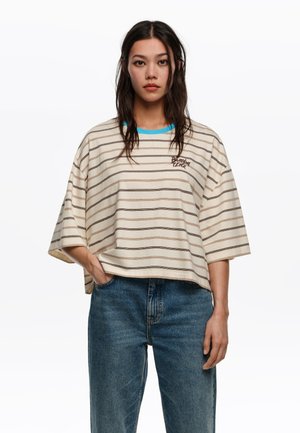 Mujer joven con cabello largo y oscuro, vistiendo una camiseta oversize beige con rayas marrones y vaqueros azules, de pie frente a un fondo blanco.