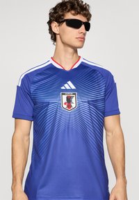 Man som bär mörka solglasögon och en blå Japans fotbollströja med vit och ljusblå randig design, stående mot en enkel ljus bakgrund.