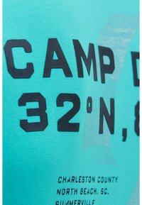 Turquoise fabric with bold black text "CAMP 32° N," and smaller text listing Charleston County, North Beach, SC, and Summerville.