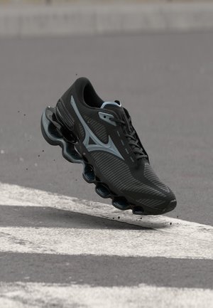Black and grey running shoe with spring sole design, mid-air above a textured asphalt surface with white road markings.