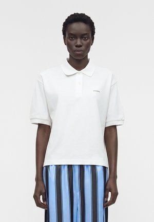 Μαύρη γυναίκα που φορά λευκό κοντομάνικο πόλο και μπλε παντελόνι με μαύρες ρίγες, στέκεται αντιμετωπίζοντας μπροστά σε απλή φόντο.