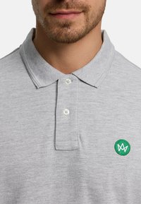 Harmaa puuvillainen poolopaita, pehmeä tekstuuri, kaksi nappia kauluksessa ja vasemmalla puolella vihreä kirjailtu logo.