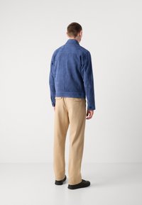 Μπλε σουέτ ζακέτα με γιακά, χωρίς ραφές και μακριά μανίκια, σε συνδυασμό με μπεζ παντελόνι και μαύρα παπούτσια, εμφανίζεται από την πίσω πλευρά.