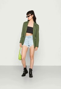 Blazer xadrez verde, top cropped preto, calções de denim claros desgastados, botins pretos, segurando uma pequena bolsa verde.