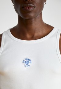 Débardeur blanc à côtes avec le texte brodé bleu "On Vacation CLUB" et un motif de soleil sur le devant. Modèle sans manches et ajusté.