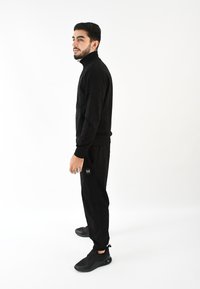 Sudadera negra con cremallera y pantalones a juego con un acabado texturizado. Presenta un pequeño logo en el pantalón y zapatillas deportivas negras.