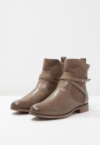 Botines marrones hechos de materiales lisos y tipo suave, con un zipper lateral, una correa decorativa y un tacón bajo de madera.