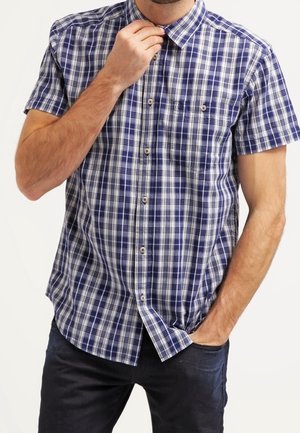 Homme portant une chemise à manches courtes à carreaux bleu et blanc, ajustant le col d'une main et l'autre main dans la poche.